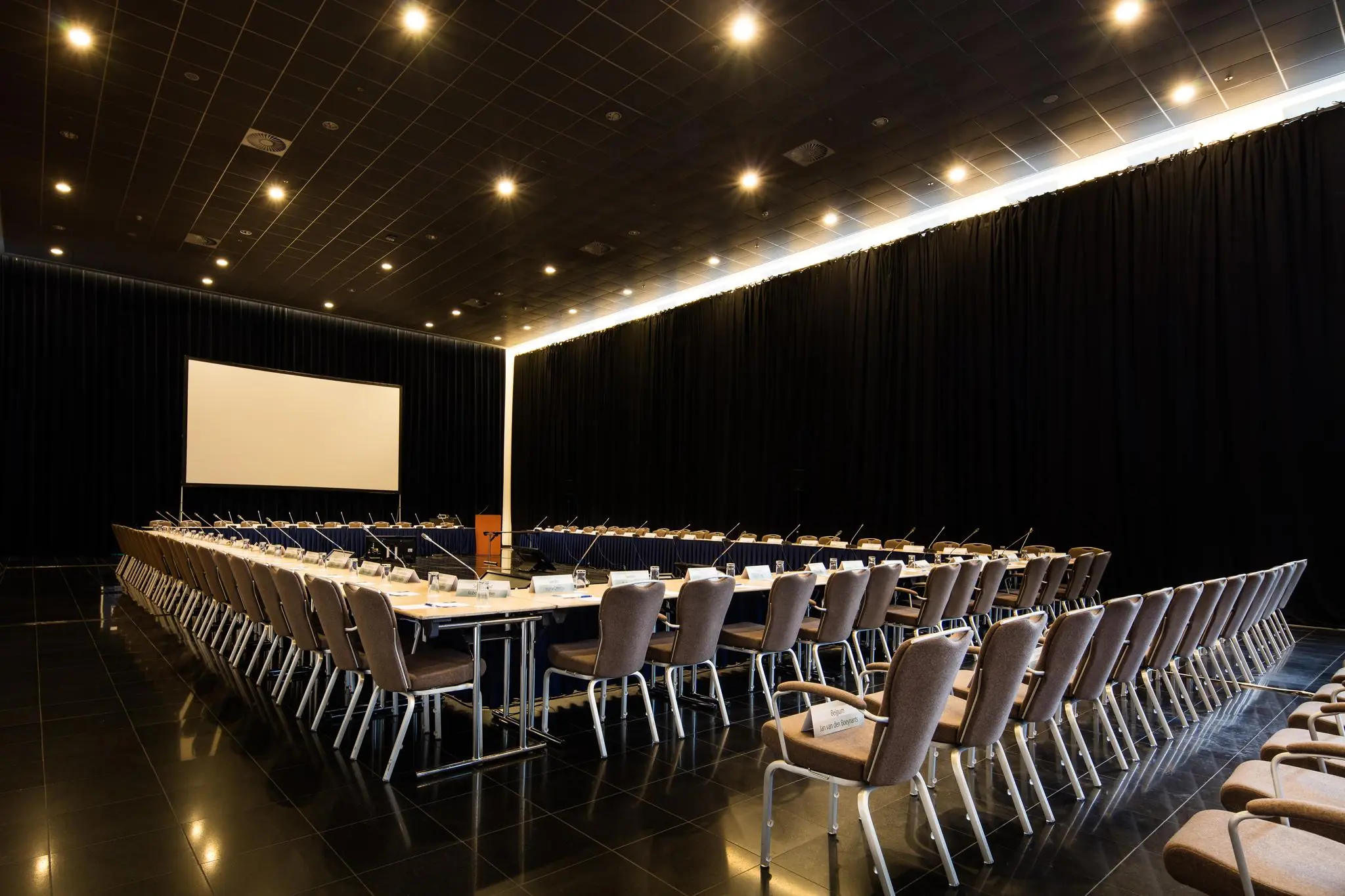 Vergaderzaal aan de Prinses Beatrixlaan met een U-vormige opstelling van stoelen en microfoons, klaar voor een conferentie of bijeenkomst.